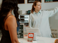 Cafe owner points at menu and pricing for customer behind counter.