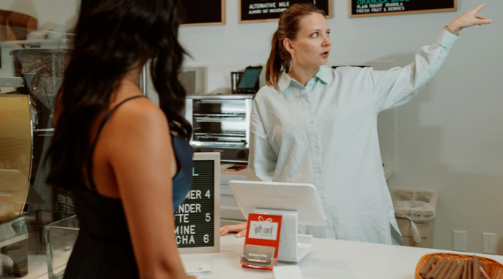 Cafe owner points at menu and pricing for customer behind counter.