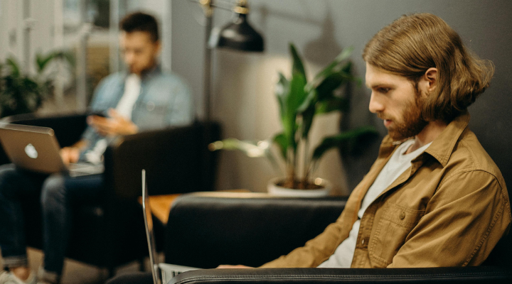 Man in brown jacket using laptop in cafe with other worker behind him.