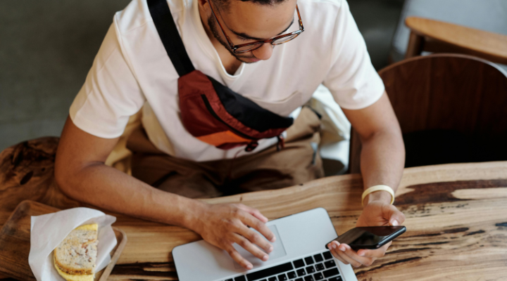 Man with a laptop and sandwich on the desk in front of him using a phone.