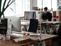 Open plan office with desks and computers, apple monitor in foreground, man working on computer in background.