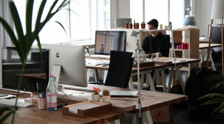 Open plan office with desks and computers, apple monitor in foreground, man working on computer in background.
