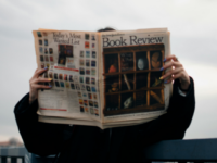 Person with coloured nails leaning against bridge rail reading the new york times book review.