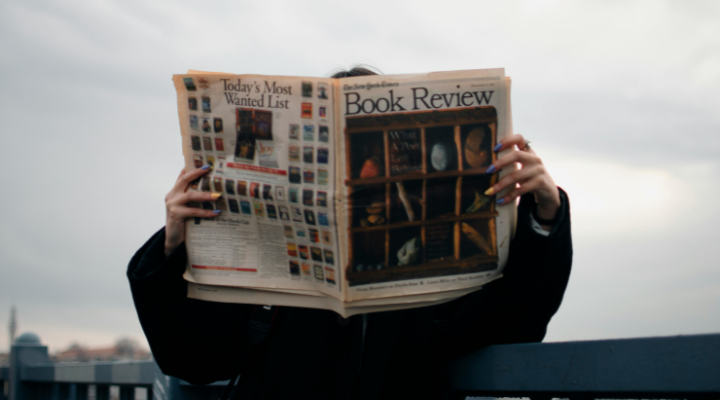 Person with coloured nails leaning against bridge rail reading the new york times book review.