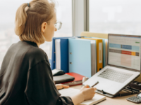 Woman sitting at desk looking at laptop with spreadsheet application open.