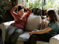 Two women sit on a couch in a sunny plant-filled room chatting